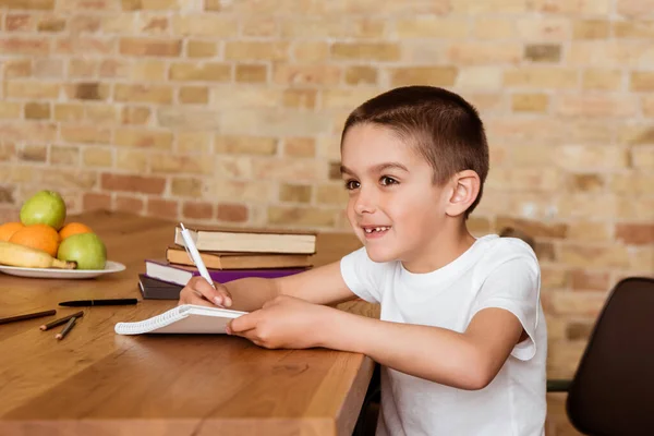 Улыбающийся ребенок пишет на блокноте рядом с книгами и фруктами на столе — стоковое фото