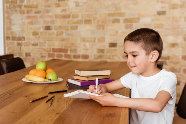 Ragazzo che scrive su copybook vicino a libri e matite sul tavolo — Foto stock