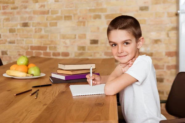 Vista laterale del bambino che guarda la fotocamera mentre scrive sul taccuino vicino a libri e frutta sul tavolo — Foto stock