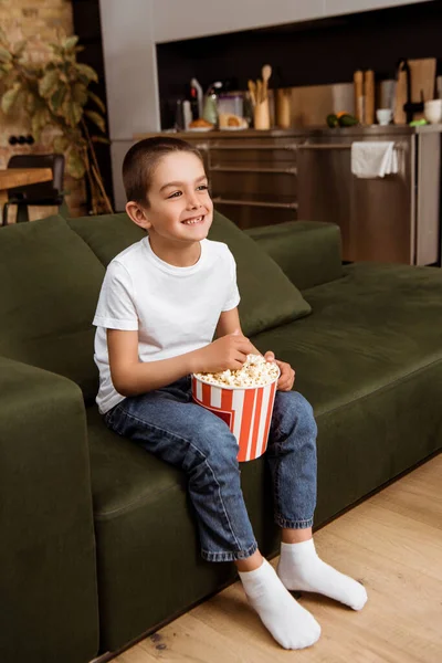 Ragazzo allegro che tiene secchio con popcorn e guarda la tv a casa — Foto stock