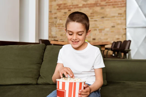 Lächelnder Junge isst Popcorn, während er im Wohnzimmer auf dem Sofa sitzt — Stockfoto