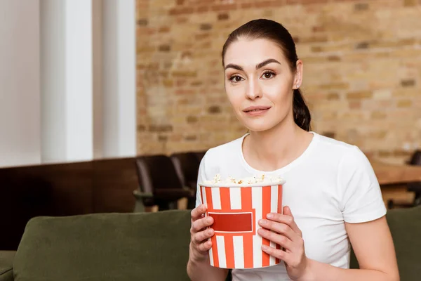 Attraktive Frau blickt in die Kamera, während sie zu Hause Eimer Popcorn auf der Couch hält — Stockfoto