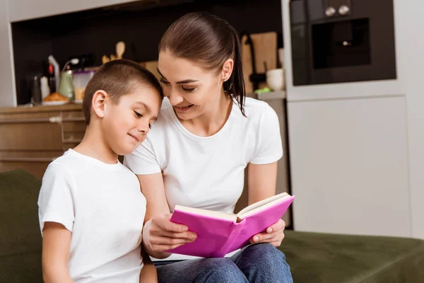 Sorridente madre tenendo libro vicino carino figlio sul divano — Foto stock