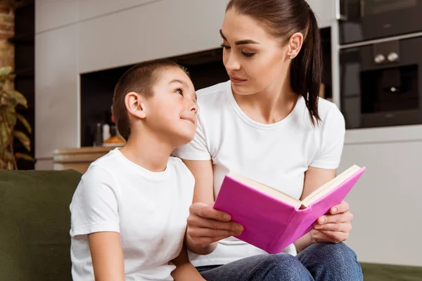 Mutter schaut süßen Sohn an, während sie Buch auf Couch im Wohnzimmer hält — Stockfoto