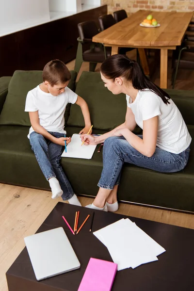 Mère et fils dessinent avec des crayons de couleur près d'un ordinateur portable et réservent sur une table basse dans le salon — Photo de stock