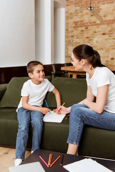 Enfoque selectivo del niño sonriente mirando a la madre mientras dibuja con lápiz de color en el sofá - foto de stock