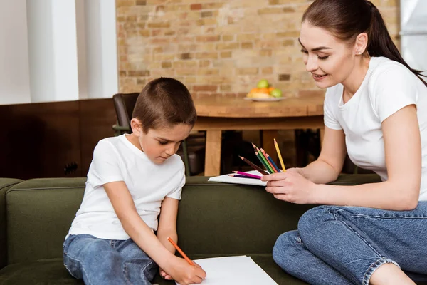 Smiling mother holding color pencils near son drawing on paper on sofa — Stock Photo