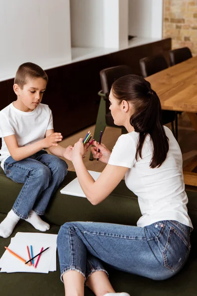 Mother holding color pencils near son and paper on couch at home — Stock Photo