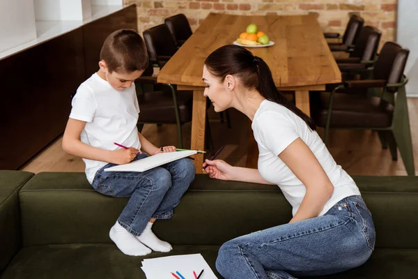 Усміхнена мати дивиться на сина, малюючи кольоровими олівцями вдома — стокове фото