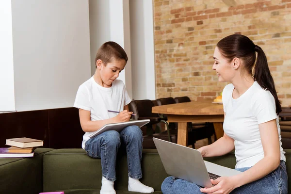 Lächelnde Mutter hält Laptop neben Sohn und zeichnet Skizzenbuch auf Couch — Stockfoto