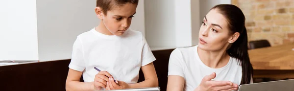 Orientación panorámica de la madre señalando con la mano a la computadora portátil al hijo sosteniendo lápiz y cuaderno de bocetos en casa - foto de stock