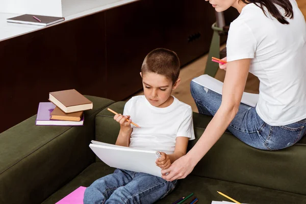 Madre sosteniendo cuaderno de bocetos mientras hijo con lápiz de color sentado cerca de libros en el sofá - foto de stock