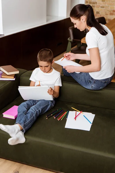 Madre y niño dibujando con lápices de color en cuaderno de bocetos cerca de libros en el sofá en casa - foto de stock