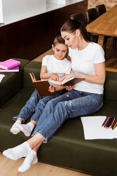 Madre e hijo lindo señalando con los dedos mientras lee libros cerca de lápices de color y papel en el sofá - foto de stock