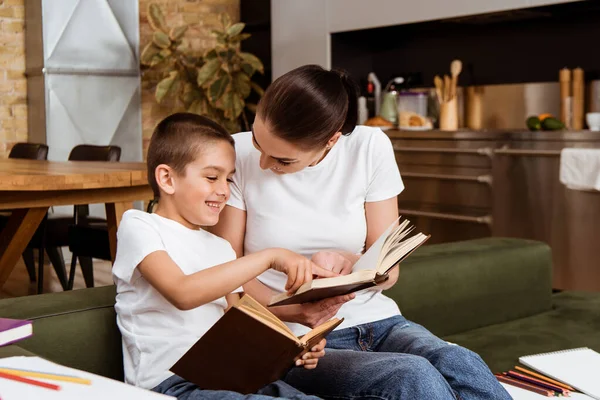 Selektiver Fokus eines lächelnden Jungen, der mit dem Finger zeigt, während er Bücher mit Mutter auf der Couch liest — Stockfoto