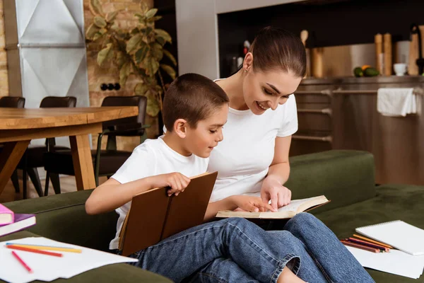 Enfoque selectivo del niño y la madre sonriente señalando con los dedos mientras lee libros cerca de lápices de color y papel en casa - foto de stock