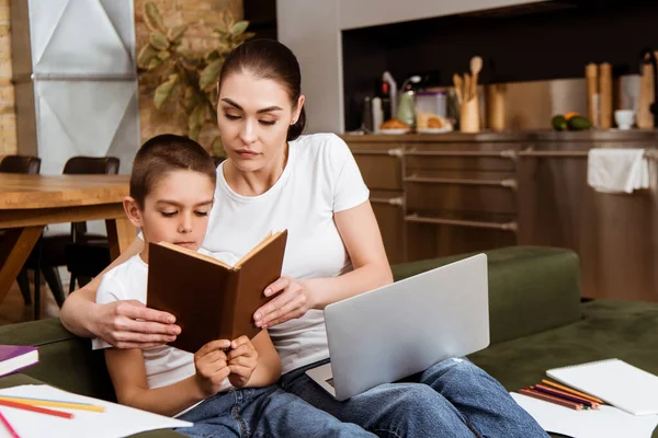 Foco seletivo de livro de leitura de mãe e criança perto de laptop, papel e lápis de cor no sofá — Fotografia de Stock