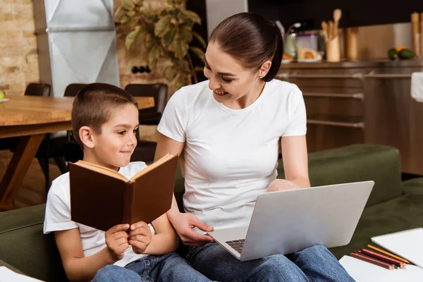 Smiling kid holding book near mother with laptop on sofa at home — Stock Photo