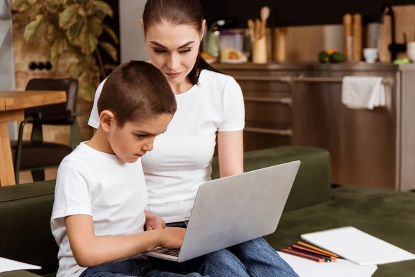 Concentration sélective du garçon en utilisant un ordinateur portable pendant l'éducation en ligne près de la mère, crayons de couleur et papier sur le canapé — Photo de stock