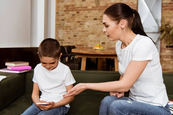 Madre señalando con la mano cerca del hijo sonriente usando el teléfono inteligente en el sofá - foto de stock