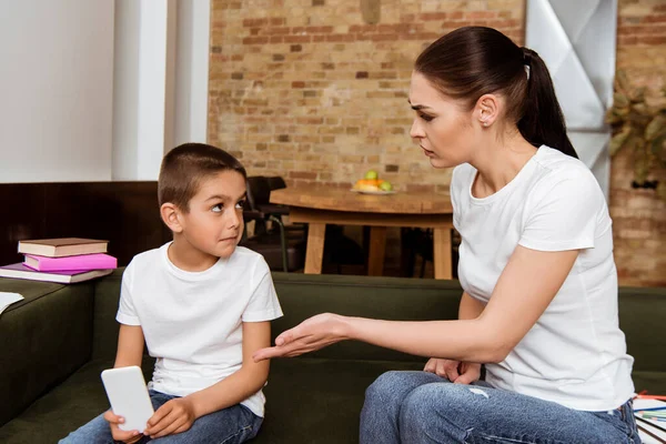 Angry mother pointing with hand at son holding smartphone on sofa — Stock Photo