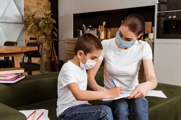 Мать и ребенок в медицинских масках рисуют цветными карандашами на бумаге рядом с книгами на диване — стоковое фото
