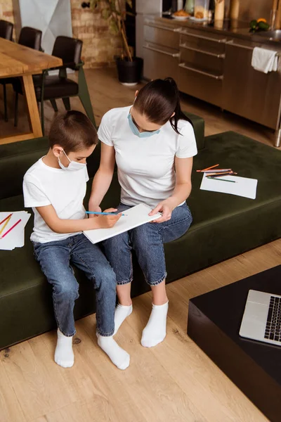 Hochwinkelaufnahme von Mutter und Sohn in medizinischen Masken Zeichnung mit Farbstiften in der Nähe von Laptop auf Couchtisch — Stockfoto