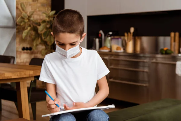 Мальчик в медицинской маске рисует на альбоме эскизов дома — стоковое фото
