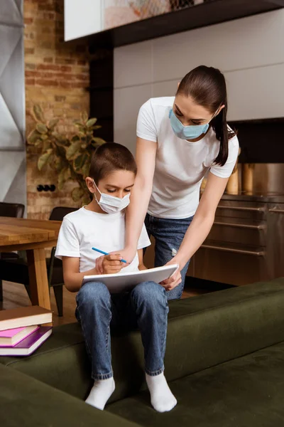 Мать в медицинской маске держит альбом эскизов рядом с сыном с карандашом и книгами на диване дома — стоковое фото