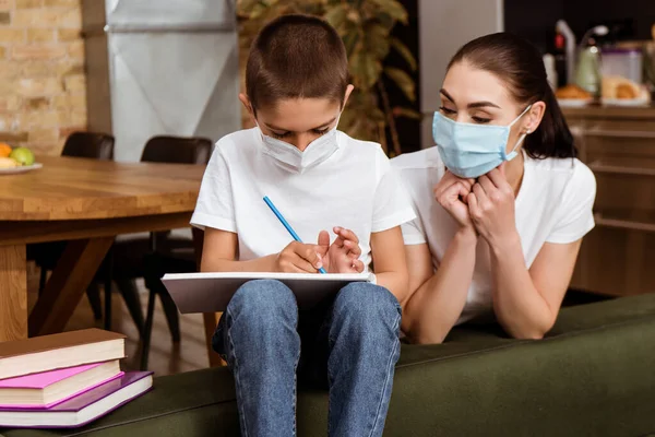 Mother in medical mask looking at kid drawing on sketchbook near books at home — Stock Photo