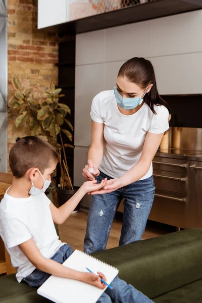 Selective focus of woman in medical mask using hand sanitizer near son holding pencil and sketchbook on couch — Stock Photo