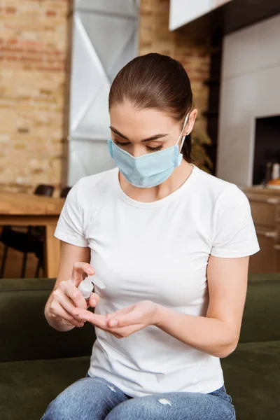 Donna in maschera medica con disinfettante per le mani sul divano a casa — Foto stock
