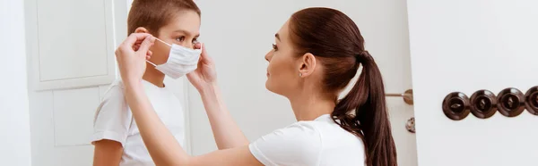 Panoramic shot of mother putting medical mask on son in hallway — Stock Photo