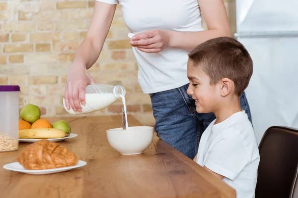 Foco seletivo da mãe derramando leite na tigela com cereais perto de filho sorridente na cozinha — Fotografia de Stock