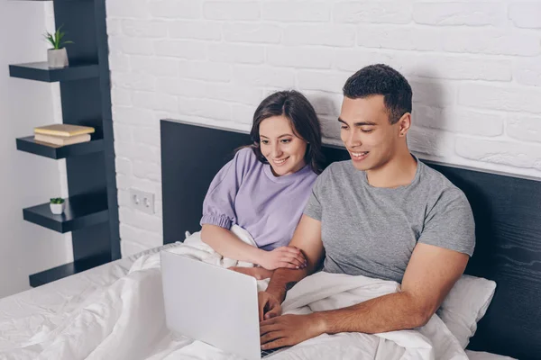 Carrera mixta freelancer utilizando portátil cerca de chica atractiva en la cama - foto de stock