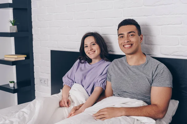 Feliz pareja interracial sonriendo en el dormitorio - foto de stock