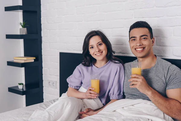 Feliz casal interracial segurando copos de suco de laranja na cama — Fotografia de Stock