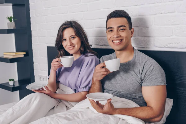 Feliz pareja interracial sosteniendo tazas de café y platillos en la cama - foto de stock