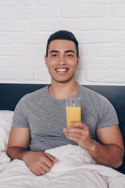 Alegre mixto raza hombre celebración de vaso de jugo de naranja en cama - foto de stock