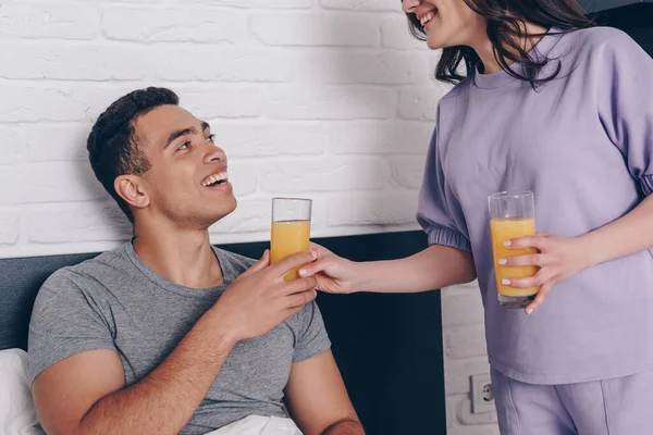 Mujer alegre dando vaso de jugo de naranja a novio de raza mixta en el dormitorio - foto de stock