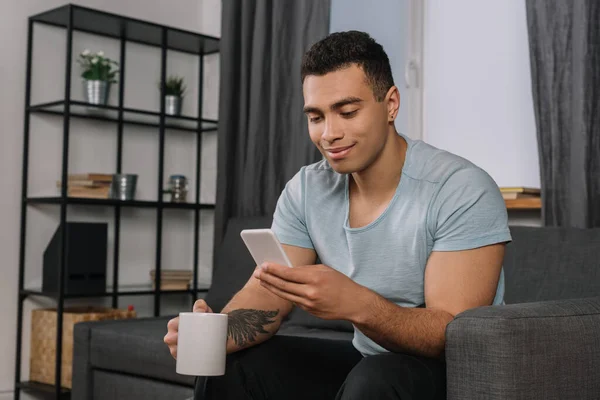 Happy mixed race man with tattoo holding cup and using smartphone — Stock Photo