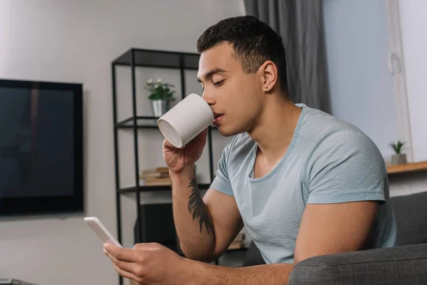 Bonito misto raça homem com tatuagem beber café e usando smartphone — Fotografia de Stock