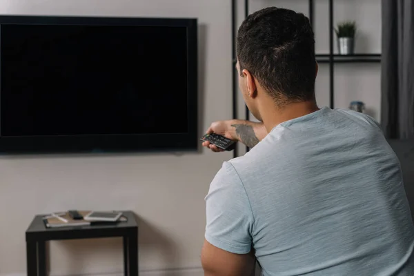 Visão traseira do homem de raça mista segurando controlador remoto perto de tv de painel plano com tela em branco — Fotografia de Stock