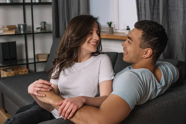 Alegre interracial casal olhando para o outro na sala de estar — Fotografia de Stock