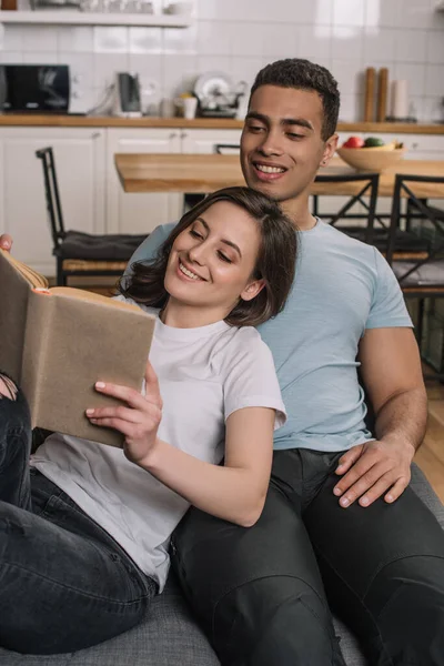 Messa a fuoco selettiva di felice donna lettura libro vicino allegro fidanzato razza mista — Foto stock