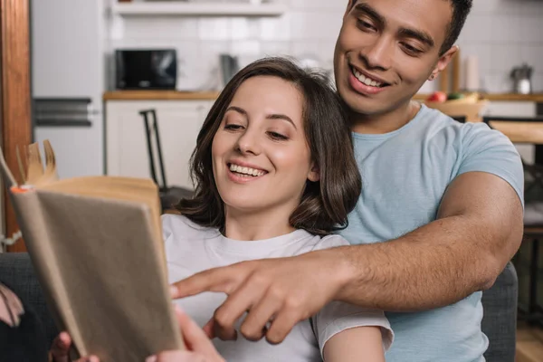 Selective focus of mixed race man pointing with finger at book near attractive girlfriend — Stock Photo