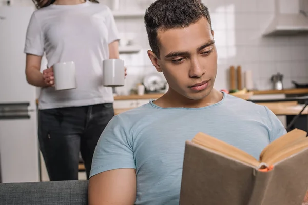 Selective focus of mixed race man reading book near girl with cups of coffee — Stock Photo