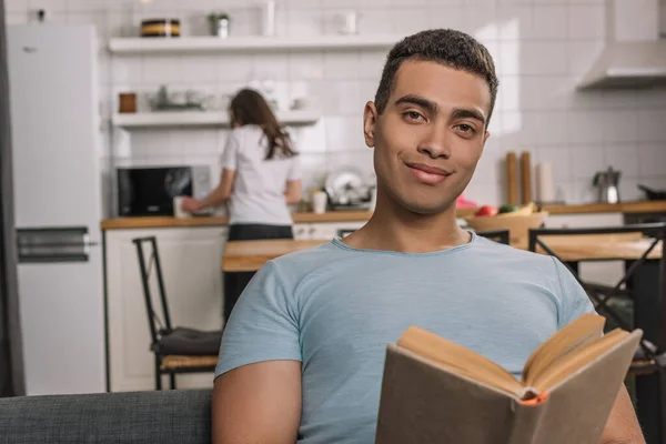 Избирательный фокус счастливого смешанного расового человека, держащего книгу дома — стоковое фото
