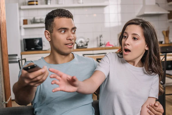 Selective focus of surprised mixed race man looking at emotional girlfriend reaching remote controller — Stock Photo