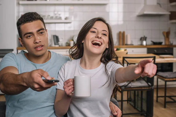 Messa a fuoco selettiva di uomo razza mista in possesso di telecomando vicino fidanzata felice ridere e puntare con il dito — Foto stock